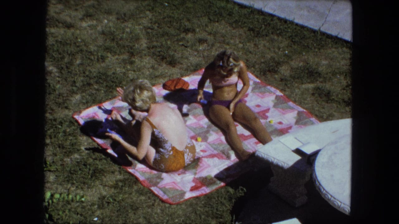 1974 Fort Wayne home-movie frame showing two adult women sunbathing on a patterned blanket beside a patio edge.