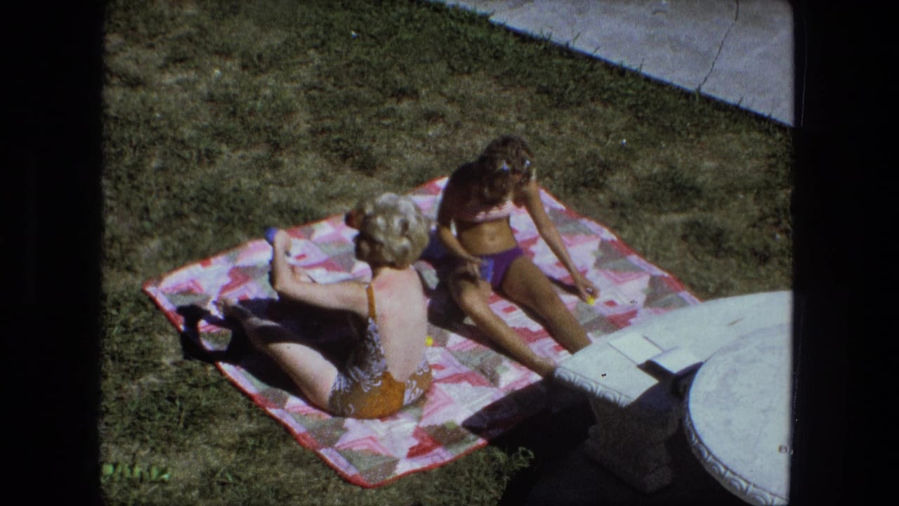 Two adult women relax on a pink blanket in a Fort Wayne backyard, filmed from above in 1974 8mm footage.