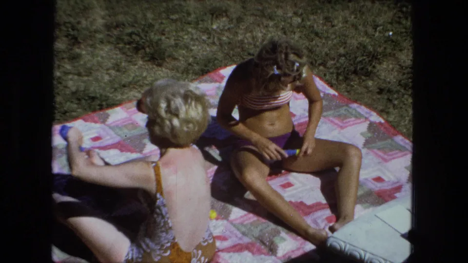 Closer 1974 home-movie view of two adult women on a sunny Fort Wayne backyard blanket, one seated facing the camera angle.