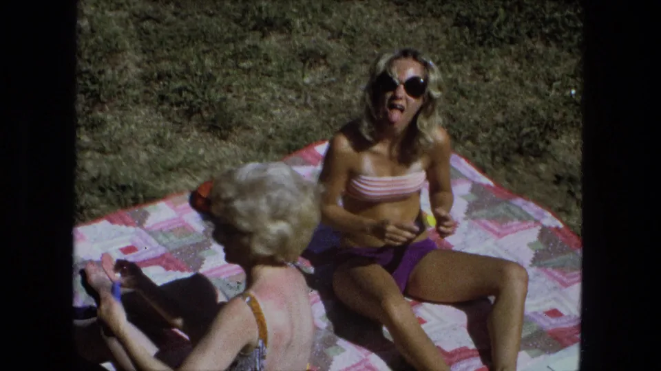 Playful 1974 Fort Wayne frame of an adult woman in sunglasses reacting to the camera while another woman sits nearby.