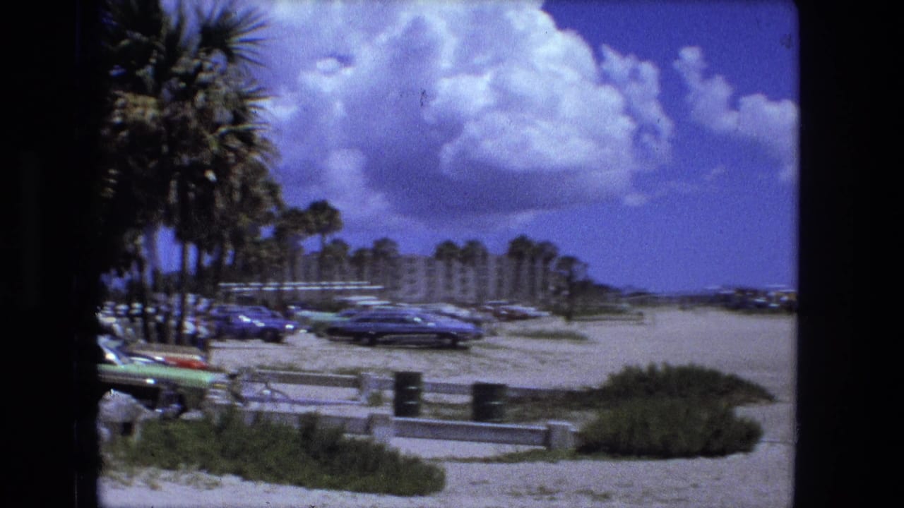 Sandy beach parking area with palm trees, cars, dunes, and clouds.