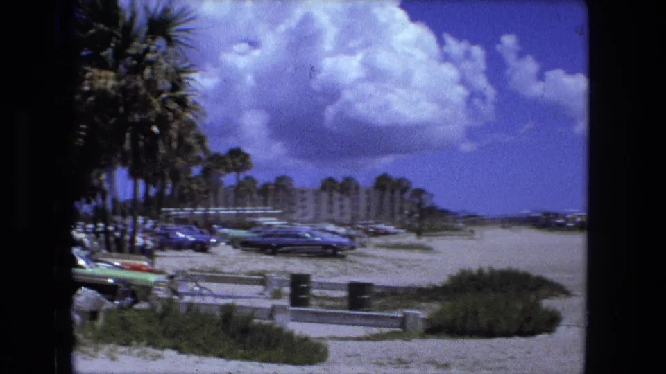 Handheld 8mm view across palm trees, parked cars, sandy beach access, dunes, and a partly cloudy coastal sky.