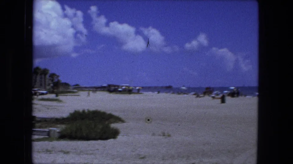 A 1970s home-movie beach view with sand, low dune grass, distant parked cars, ocean horizon, and a wide blue sky.