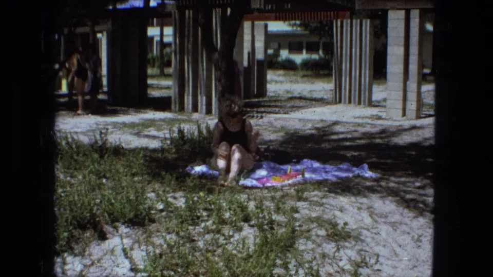 A woman in a dark swimsuit sits on a towel in shade beneath raised beachfront structures, with sand and beach grass around her.