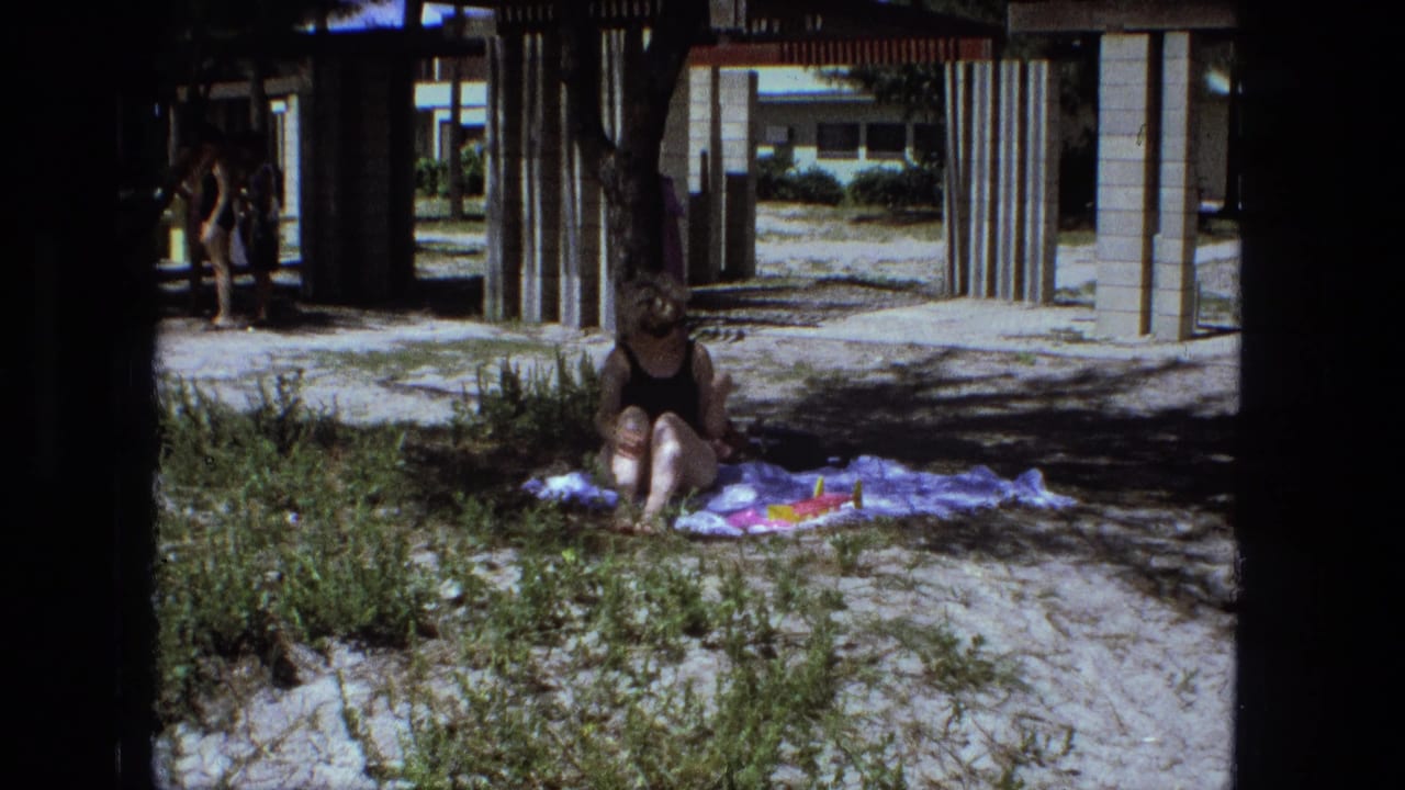 Woman in a black swimsuit sits on a colorful blanket under shade near a sunlit outdoor pool area.