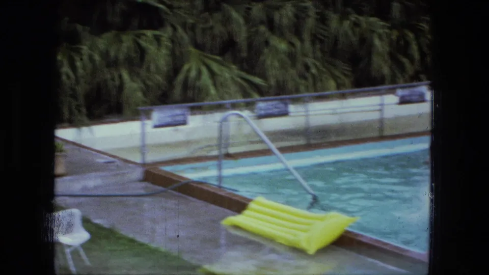 The pool deck shows a ladder, white plastic chair, yellow inflatable raft, and surrounding greenery in 1970s home-movie color.