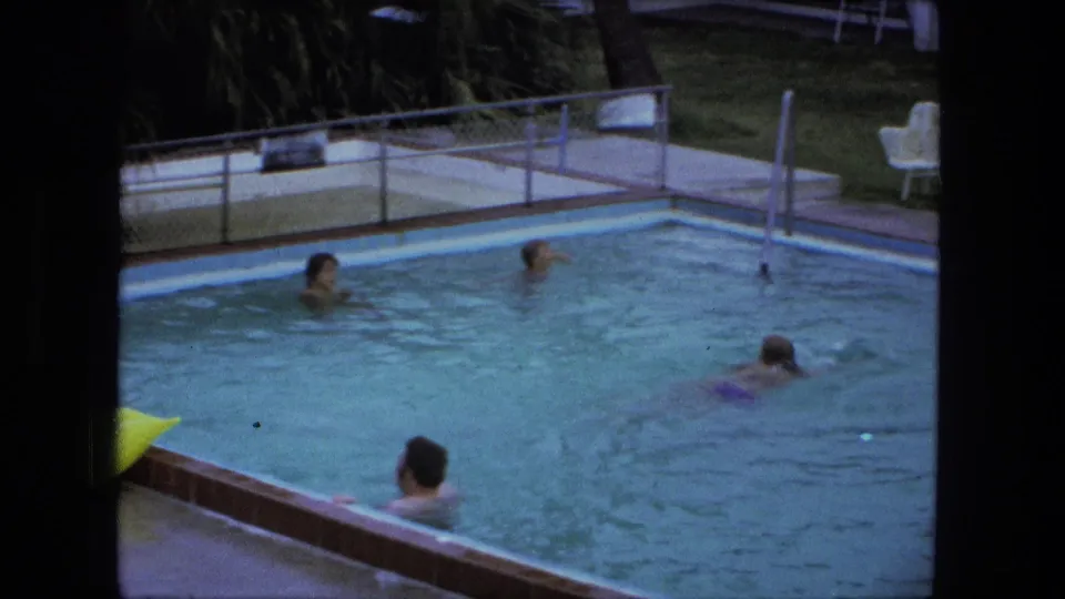 Several swimmers are visible in the outdoor pool, framed by deck furniture, railings, and greenery in the Fort Wayne home movie.