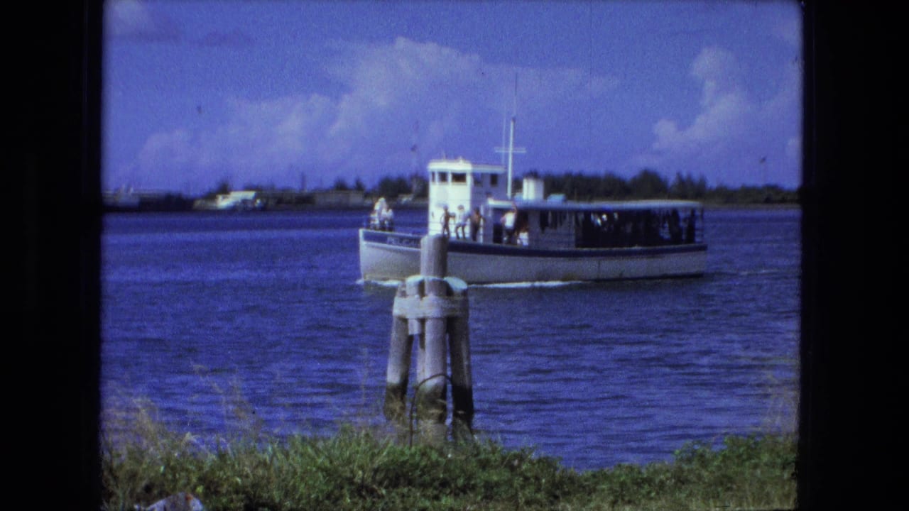 White passenger boat moving across blue river water behind wooden pilings.