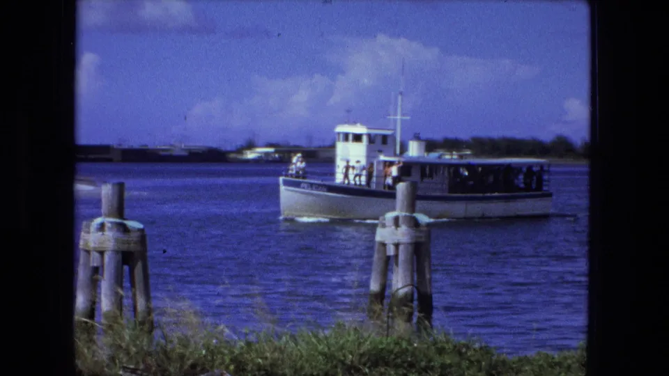The boat continues along the Fort Wayne river scene, carrying visible passengers under a covered deck near foreground pilings.