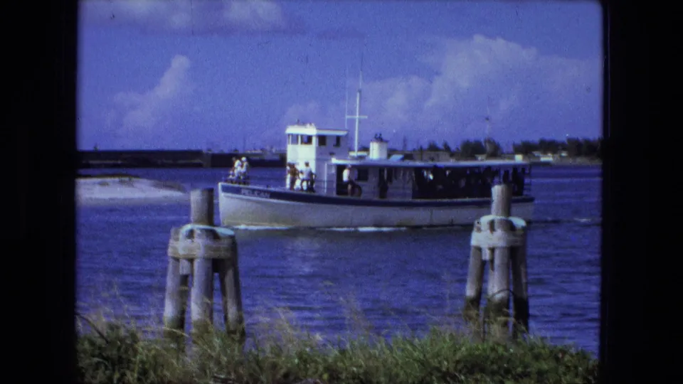 The 1974 Fort Wayne river view shows the passenger boat close to a foreground piling, with a grassy bank and distant shoreline behind it.