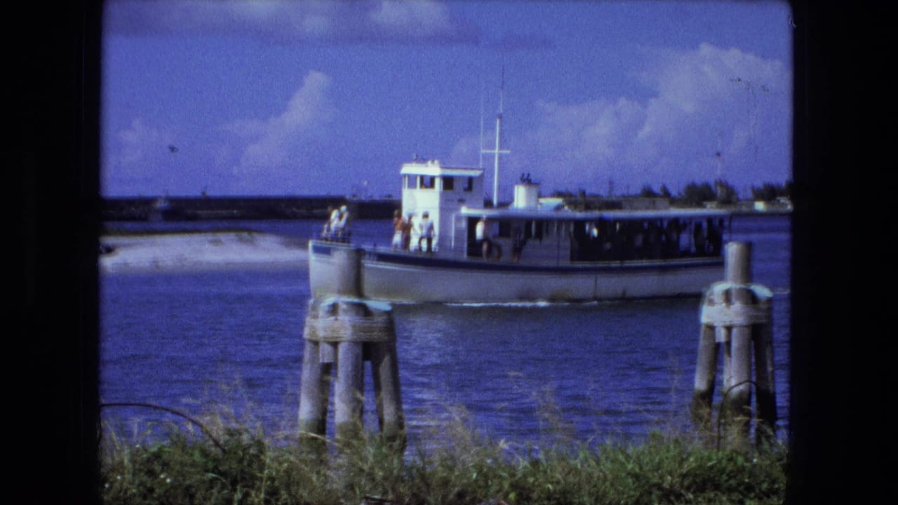 Small white passenger boat crossing blue water behind wooden pilings.