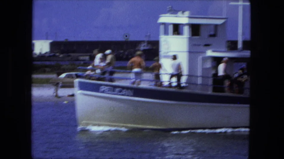 Closer 8mm view of the PELICAN passenger boat, with people standing and sitting on deck as it moves across the water.