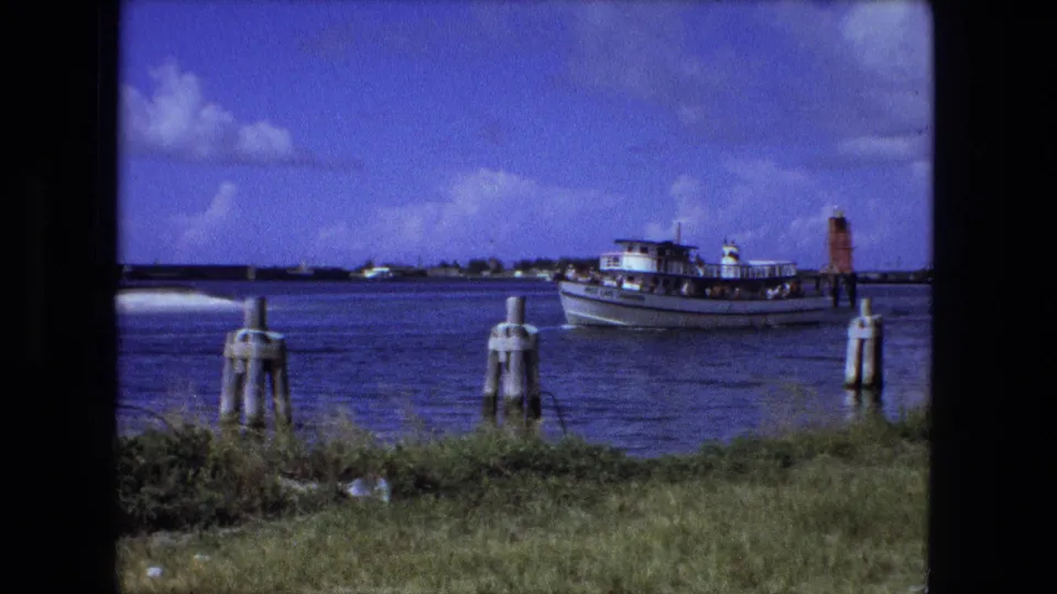 A small passenger ferry is seen farther from shore, framed by grass, wooden pilings, blue water, and a bright clouded sky.