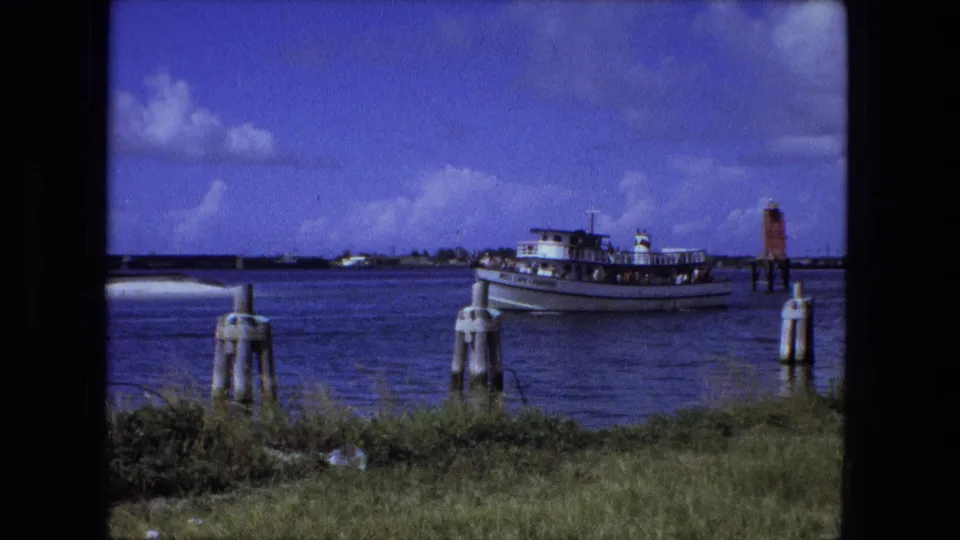 The passenger ferry moves across open water in a wide 1974 Fort Wayne home-movie view with pilings and shoreline grass in front.