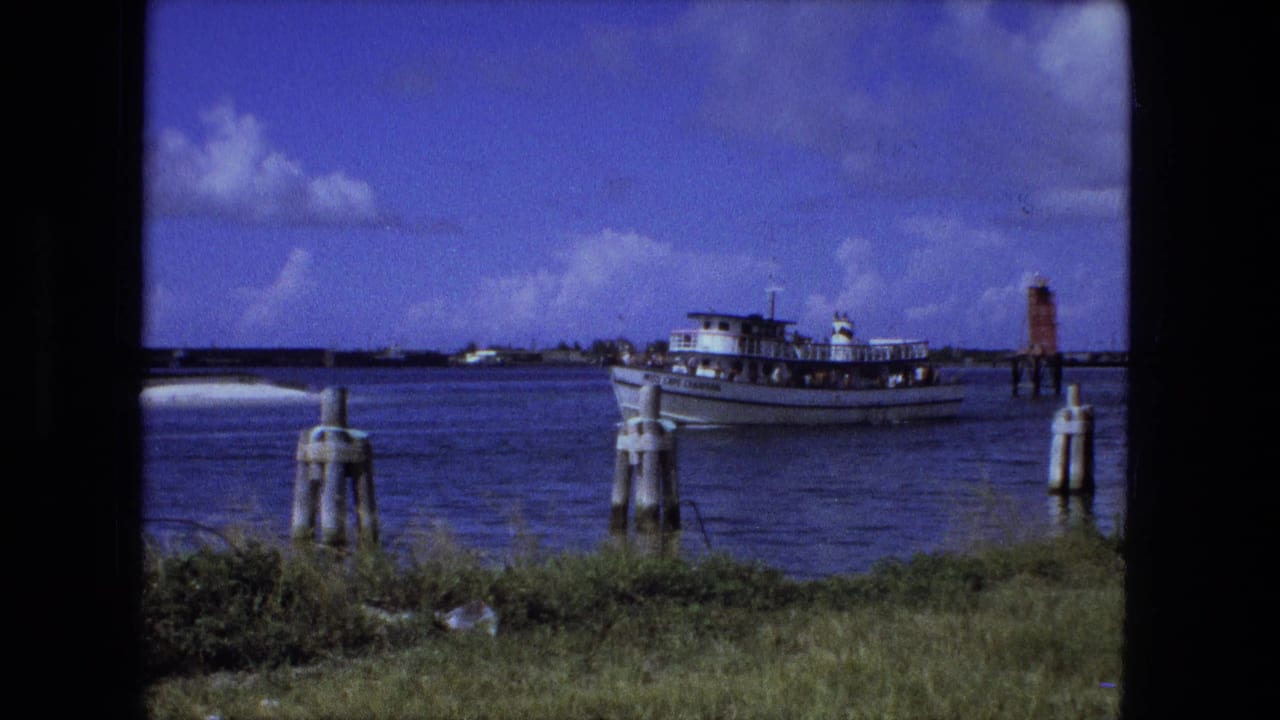 White passenger boat moving on blue water near pilings and a small lighthouse-like structure.