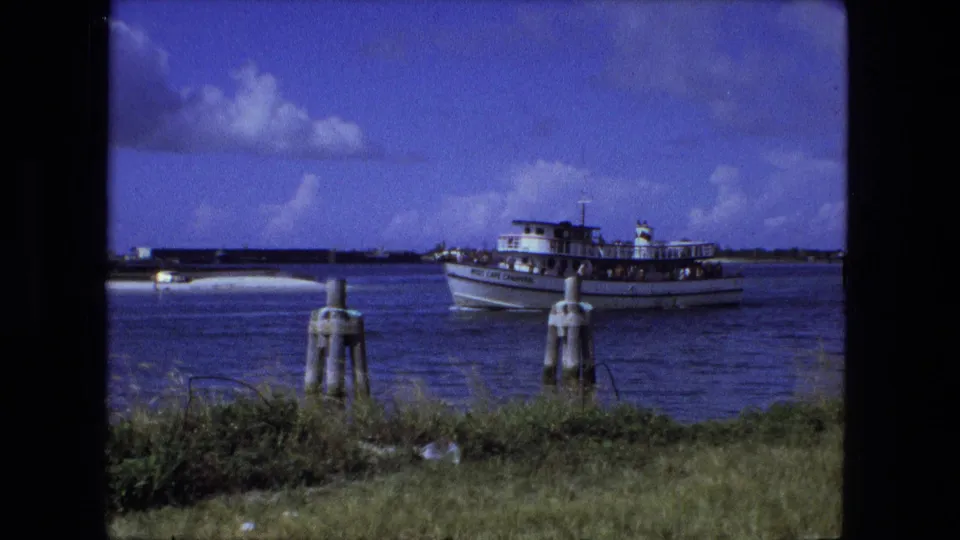 A 1970s 8mm frame shows the passenger boat mid-crossing, partly aligned behind shoreline pilings and foreground grass.
