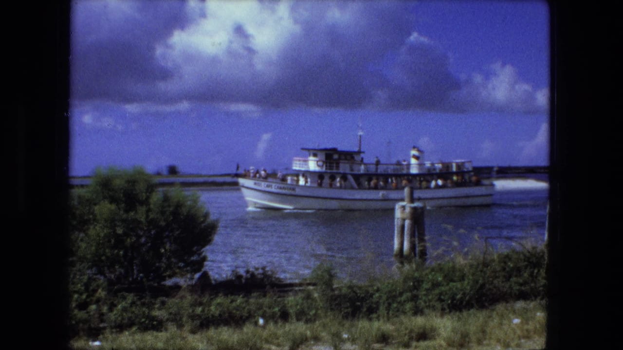 The ferry continues along the waterway, with passengers visible on deck and green shoreline vegetation in the foreground.