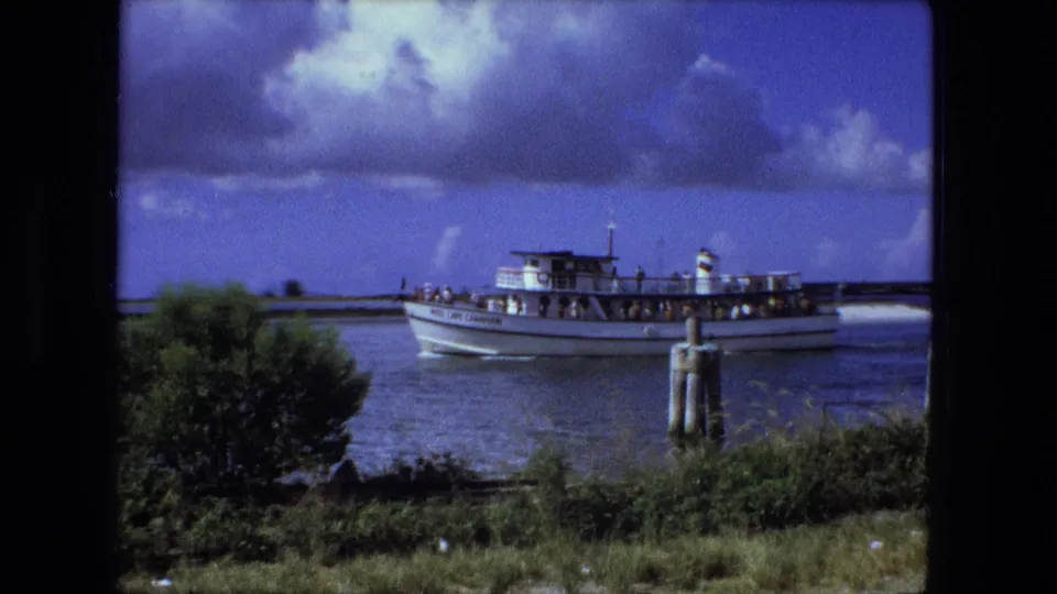 The ferry continues along the waterway, with passengers visible on deck and green shoreline vegetation in the foreground.