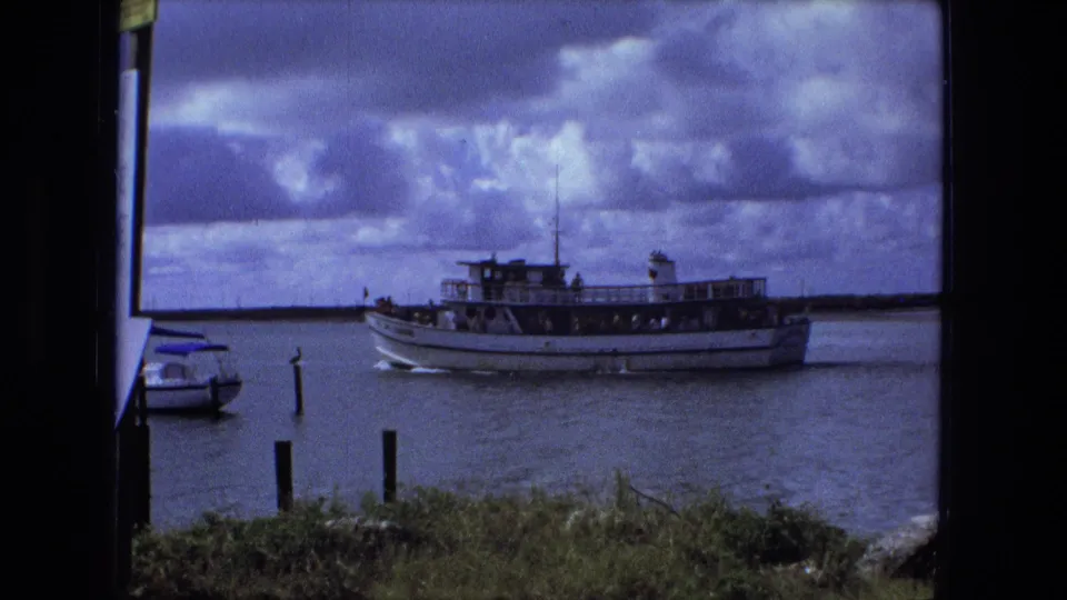 A white passenger ferry crosses the waterway near small boats, dock posts, and a grassy shoreline in the 1974 home-movie scene.