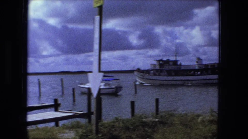 The ferry moves partly out of view near dock posts and a small boat, with a flag or signpost in the foreground.