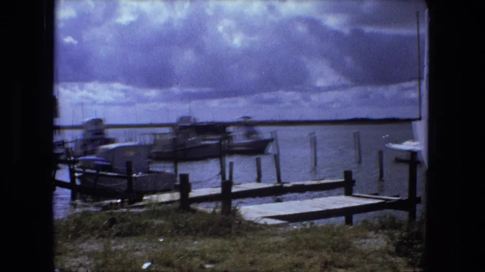 1974 Fort Wayne waterfront scene with moored boats, dock posts, calm water, and heavy cloud cover in an 8mm frame.