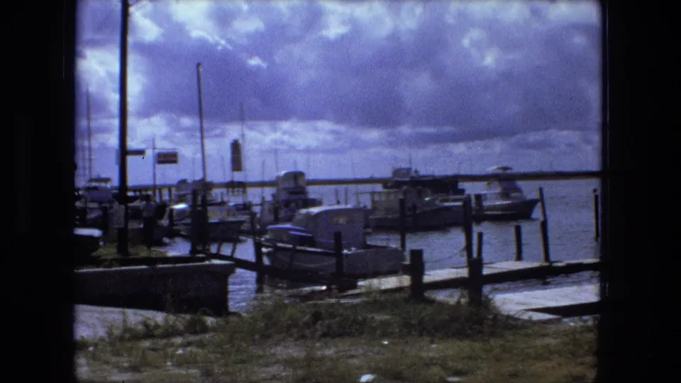 1974 Fort Wayne marina frame showing boats at docks, vertical poles, grassy shoreline, water, and cloud-filled sky.