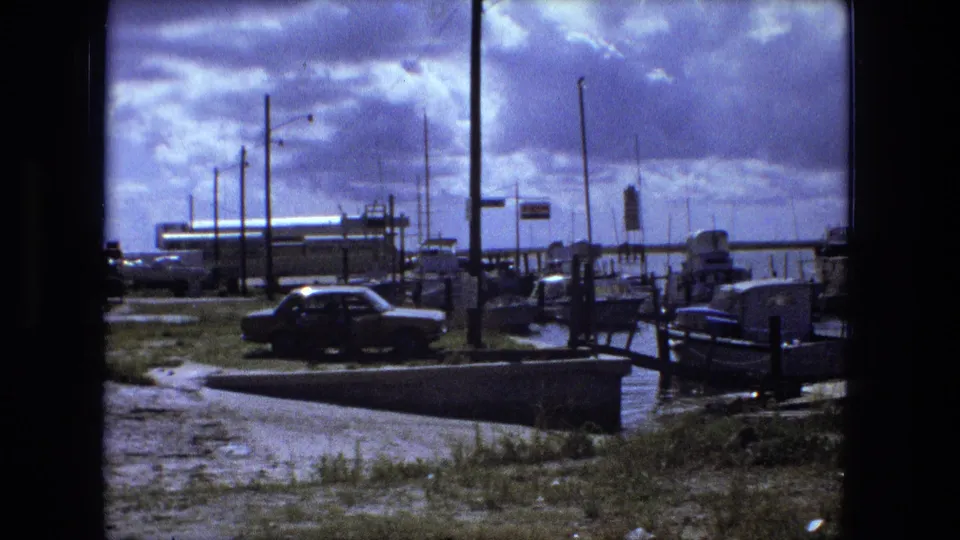 1974 Fort Wayne marina view with a parked classic car near docked boats, grassy ground, water, poles, and a cloudy sky.