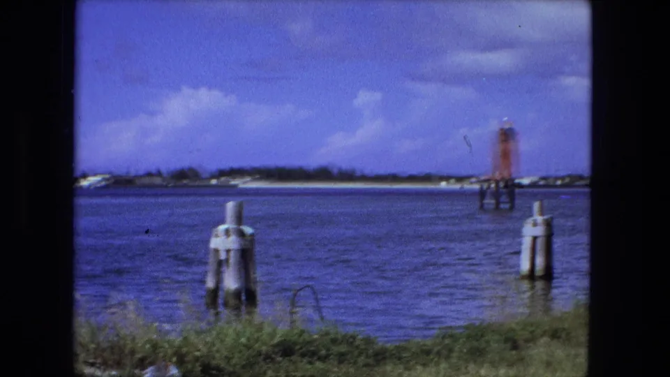 1974 Fort Wayne waterfront view with rippling water, pilings in the foreground, a red marker in the channel, and low shoreline under clouds.