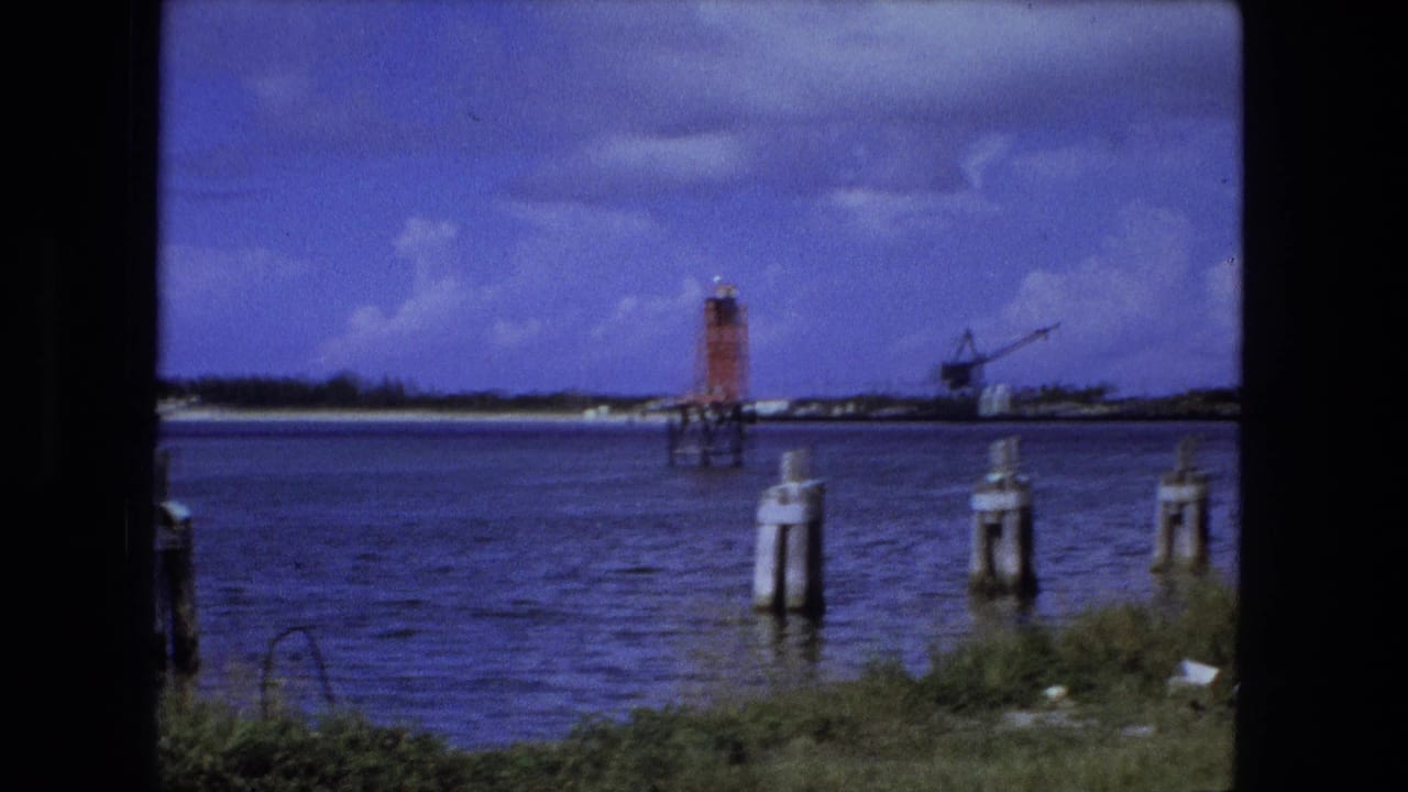 Fort Wayne 1974 waterfront frame showing pilings in the water, a red channel marker, distant industrial crane, and broad blue sky.