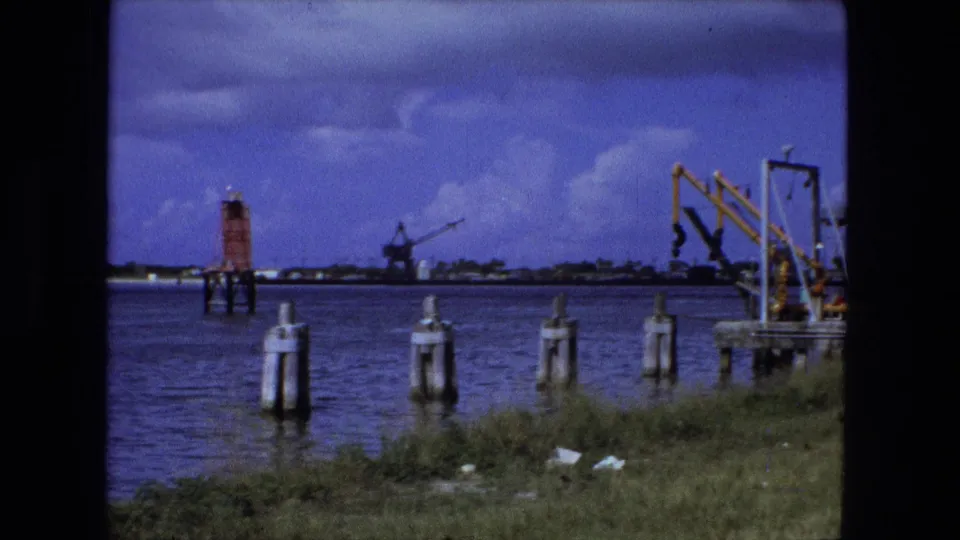 1974 home-movie frame of a Fort Wayne waterside scene with pilings, red marker, distant crane, and yellow dockside equipment.