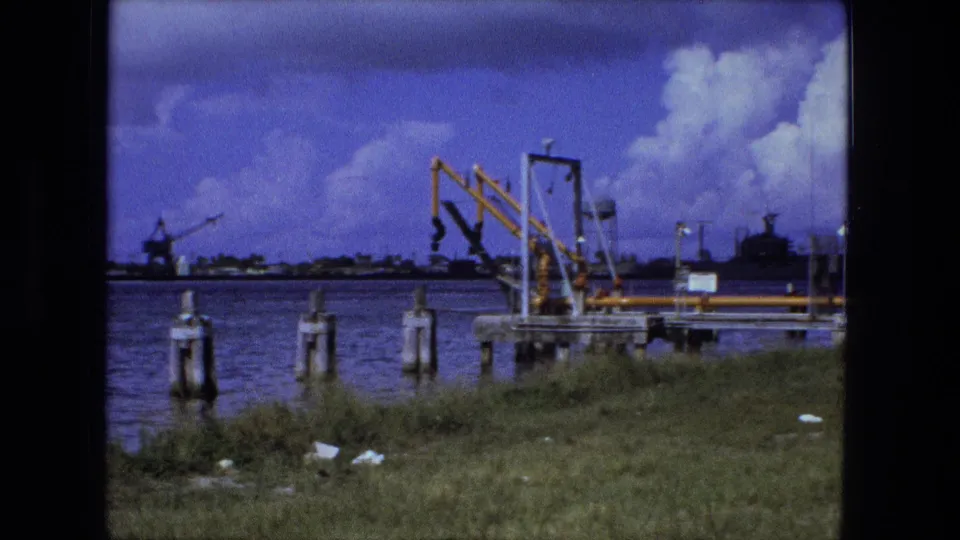 Fort Wayne 1974 waterfront view with foreground grass, pilings in the water, yellow gantry crane, distant structures, and clouded sky.
