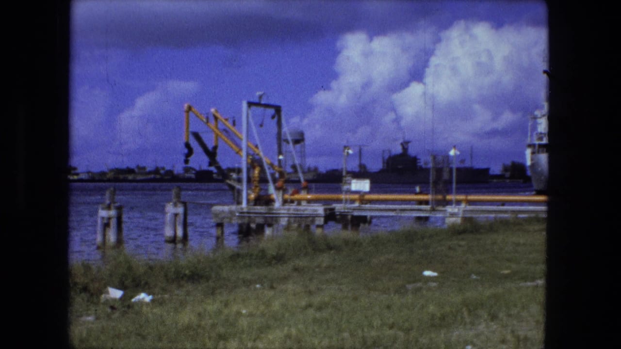 1974 8mm view, per clip metadata, of an industrial waterfront with yellow dock equipment, grassy foreground, water, and a partly visible white vessel.