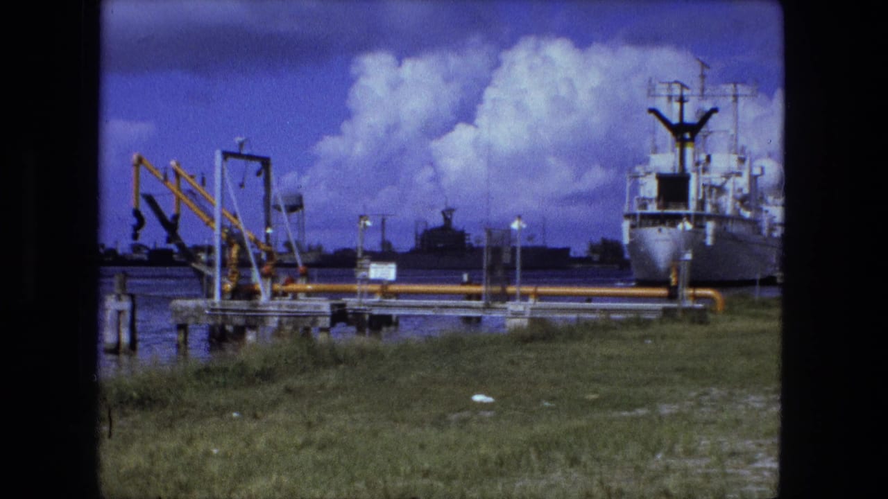Metadata-supported 1974 waterfront frame showing a white docked vessel, yellow pipework or cranes, water, grass, and distant industrial structures.