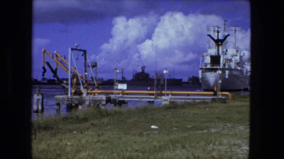 Metadata-supported 1974 waterfront frame showing a white docked vessel, yellow pipework or cranes, water, grass, and distant industrial structures.