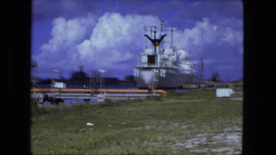 1974 8mm frame, based on clip metadata, with a docked white vessel, yellow industrial piping, waterfront grass, and hazy port structures.