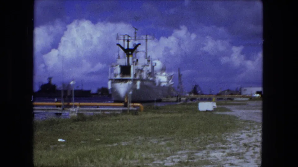 End frame from the 1974 8mm clip showing the docked vessel, yellow dock infrastructure, grass, paved ground, and distant industrial buildings.