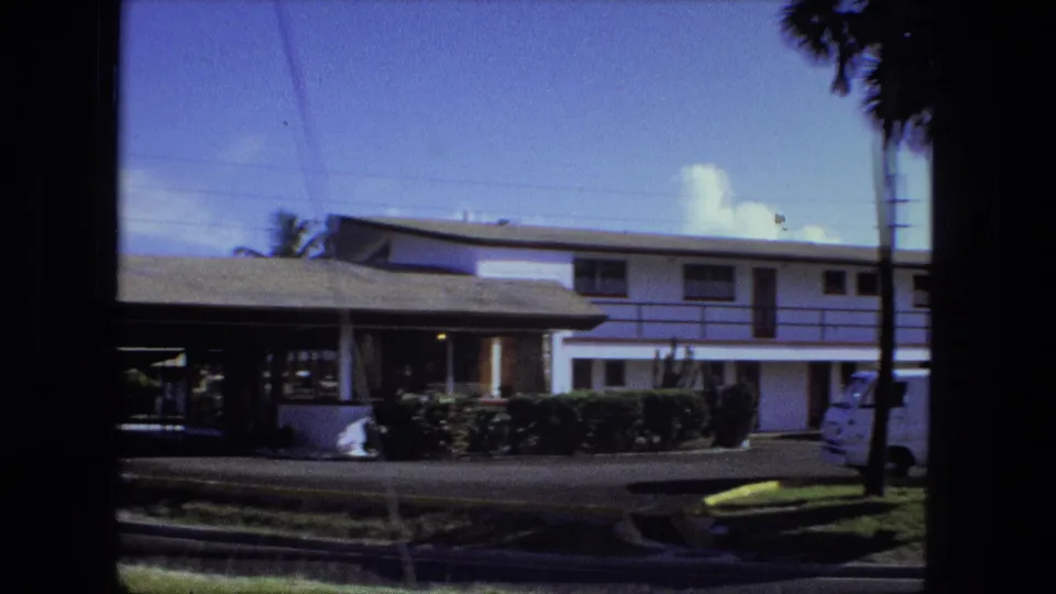 1974 Fort Wayne home-movie view of the motel's two-story guest wing, hedges, palm tree, curb, and partial roadside signage.