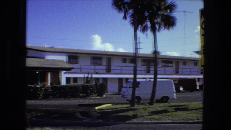 1974 Fort Wayne frame showing the motel guest wing, palm trees, hedges, roadside curb, utility poles, and a white van parked nearby.