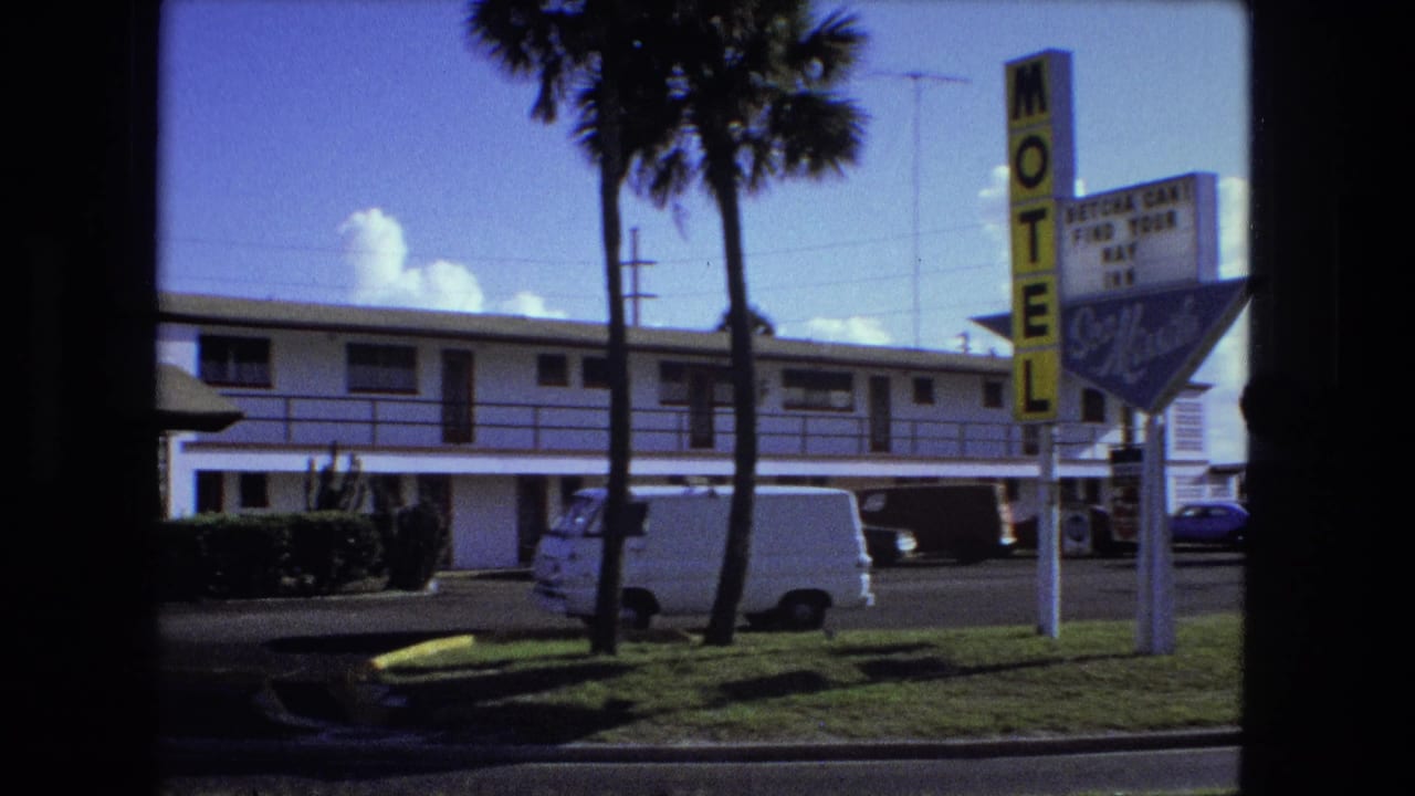 1974 home-movie frame tied by metadata to Fort Wayne, showing a roadside motel, yellow sign, palm trees, and parked vehicles.