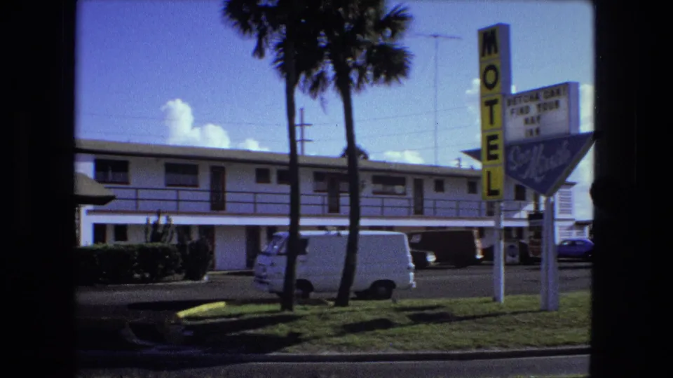 1974 home-movie frame tied by metadata to Fort Wayne, showing a roadside motel, yellow sign, palm trees, and parked vehicles.