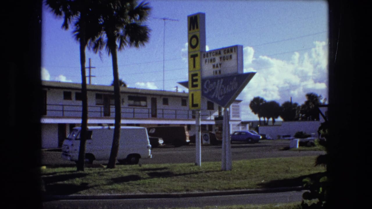Roadside motel exterior in 1974 8mm footage, with palm trees, a vertical sign, parked vehicles, and bright sky.