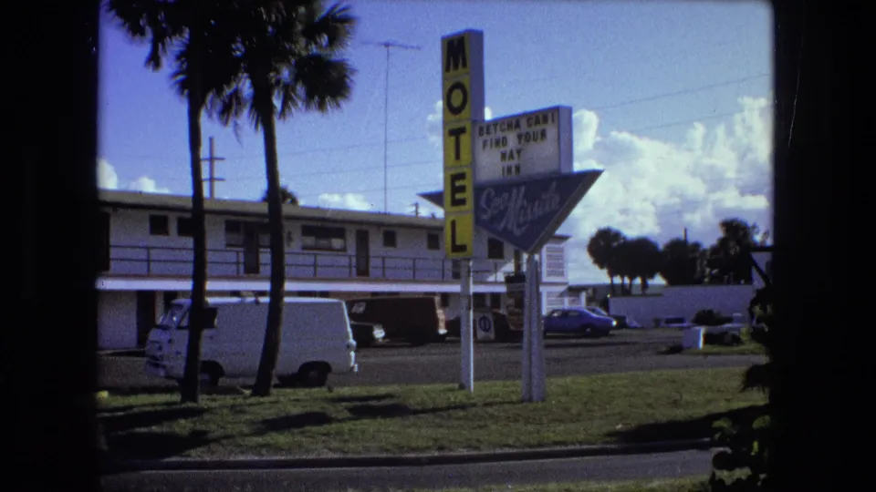 Roadside motel exterior in 1974 8mm footage, with palm trees, a vertical sign, parked vehicles, and bright sky.
