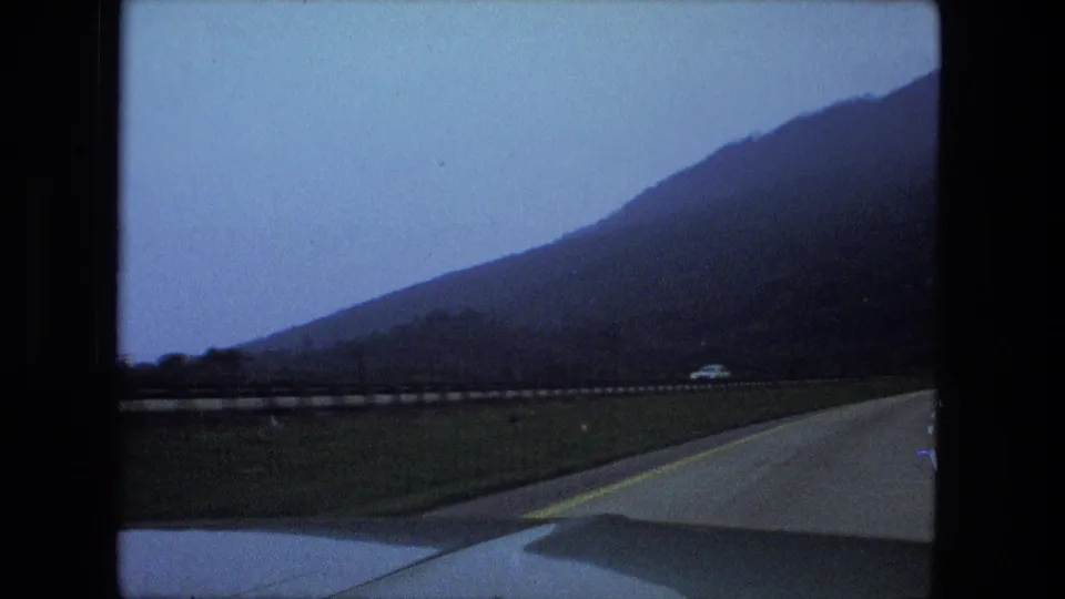 In-car 8mm travel view showing a highway, car hood, roadside barrier, open land, and a tree-covered hillside.