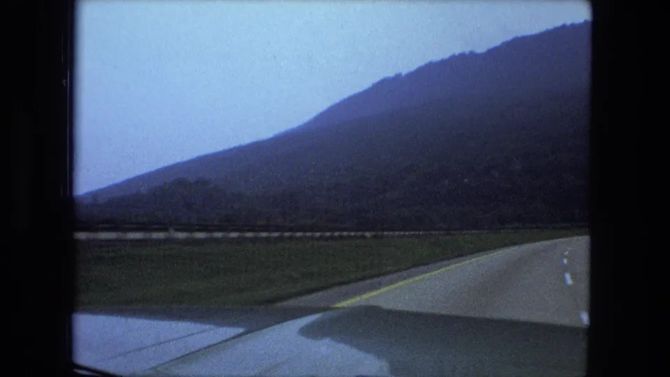 Home-movie driving view from a car, with the road curving beside open ground and a dark hillside under pale sky.