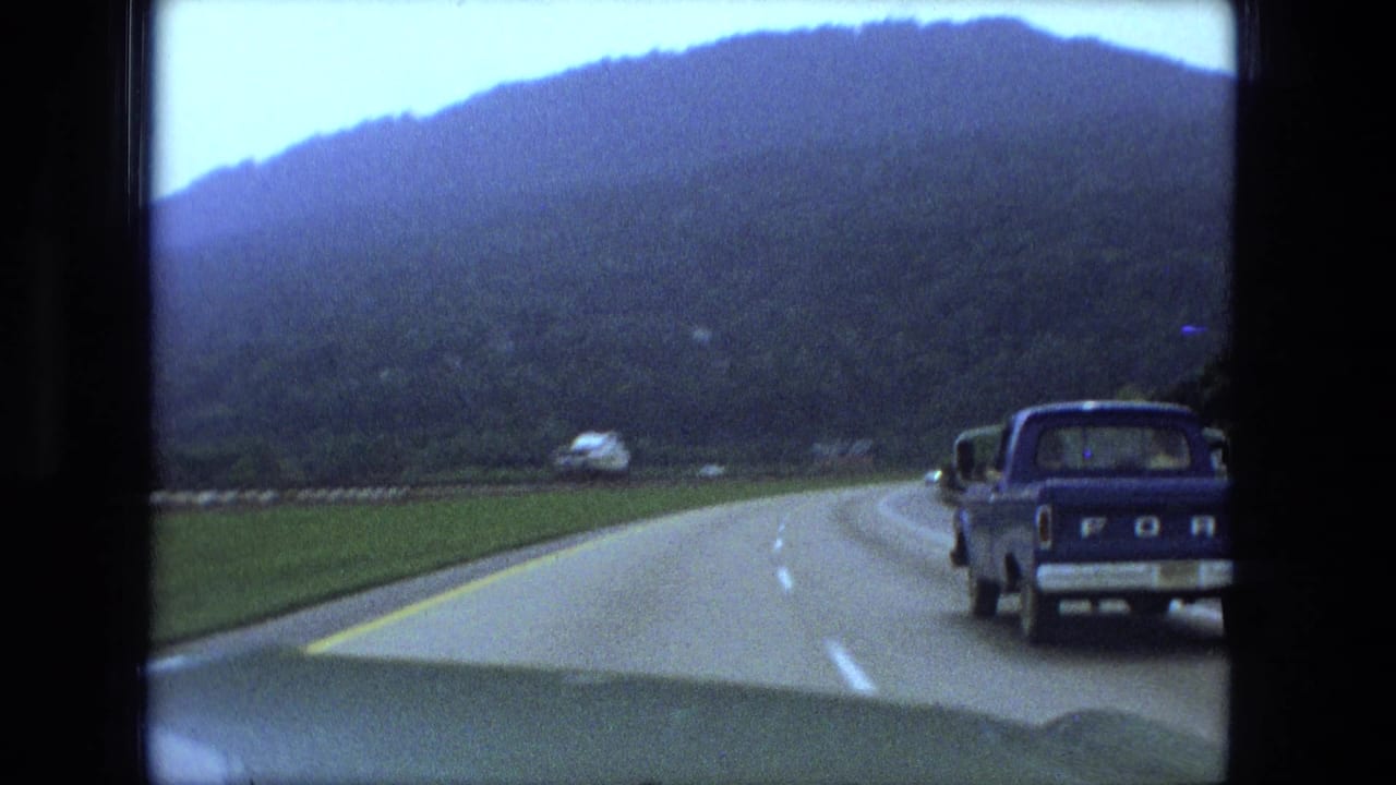 Blue Ford pickup appears beside the camera car on a 1974 home-movie highway shot with wooded hills ahead.
