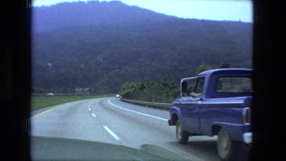 The blue Ford pickup draws alongside the moving car in 1974 8mm footage, with highway lanes and wooded hills visible.