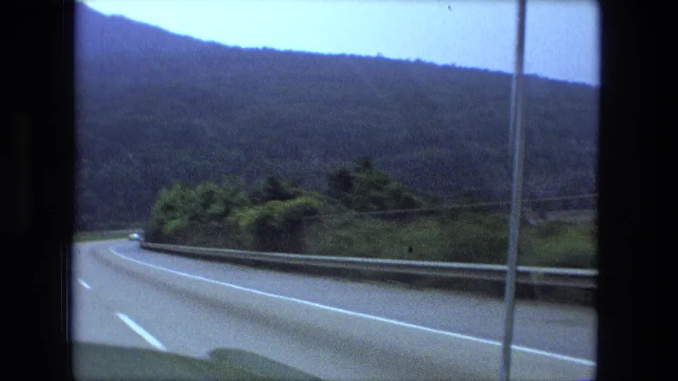 A 1974 8mm car-window view shows the highway curve continuing past guardrail and wooded hillside after the truck passes.