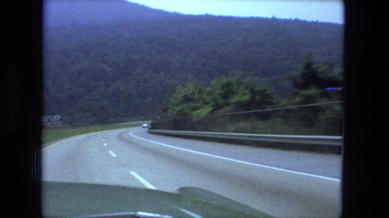 Curving highway through wooded hills seen over a car hood and windshield edge.