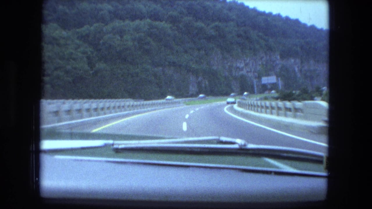 Car-window view of a divided highway with a concrete barrier, forested hillside, overhead sign, and traffic visible ahead.