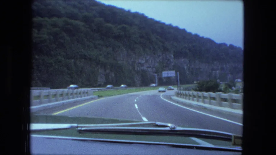 1974 8mm road view from inside a car, showing a highway curve, rocky roadside cut, trees, barriers, and vehicles ahead.